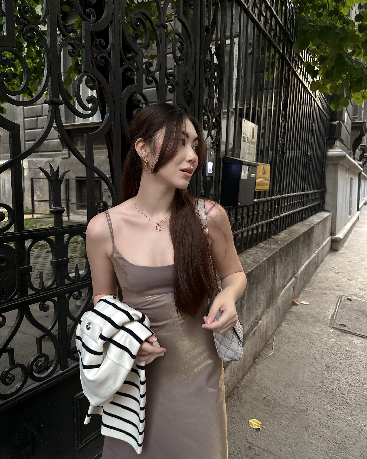 На тротуаре у черной ограды женщина в тауповом атласном платье держит сложенную полосатую вещь.